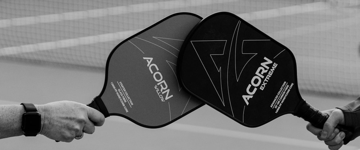 Two Acorn Pickleball paddles held by two hands, in front of a net 